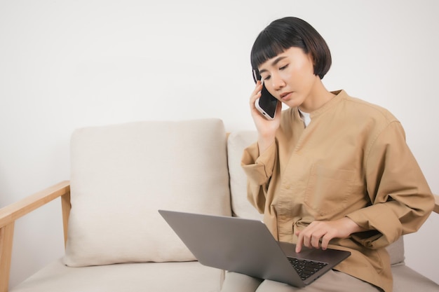 Asian woman using laptop while sitting on the sofa at home and speaking on the phone