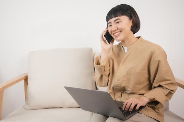 Asian woman using laptop while sitting on the sofa at home and speaking on the phone