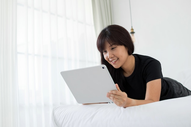 Asian woman using tablet for video conversation while relaxing on bed in home. 