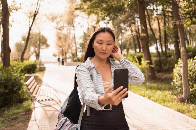 Asian woman walking in city park in summer and doing sefie on phone