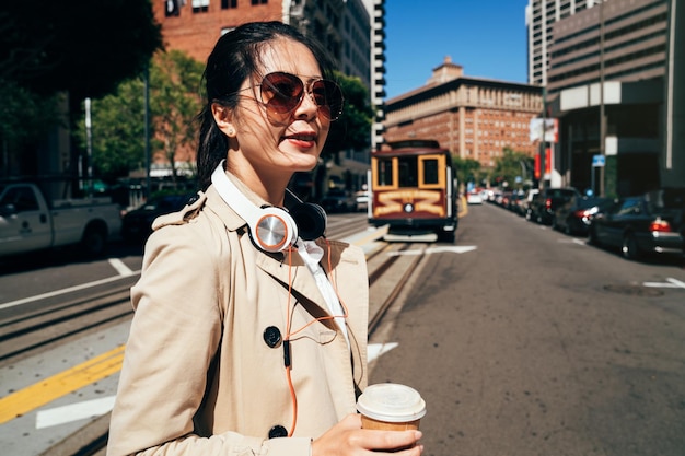 asian woman walking outdoors street hol coffee cup wearing earphones crossing road cable car streetcar railway. young college girl commute enjoy music headphones. local student going to university