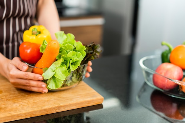 Asian woman wearing apron preparing a healthy salad with vegetables in a glass bowl.