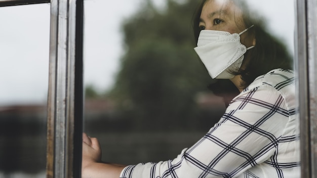 Asian woman wearing a medical mask looking out the window from her house, her eyes sad,working from home, quarantine.