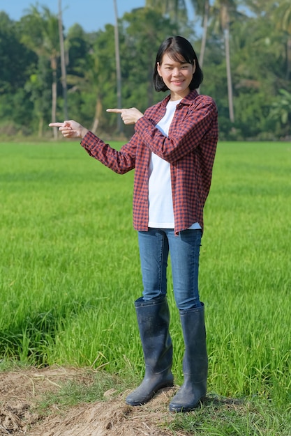 An Asian woman wearing a red striped shirt poses pointing her finger to the side in a field outdoors.