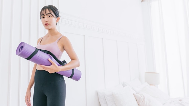 Asian woman wearing sportswear and yoga pants carrying a rolledup yoga mat standing in front of white wall background with sunlight from the window Halflength image with copy space