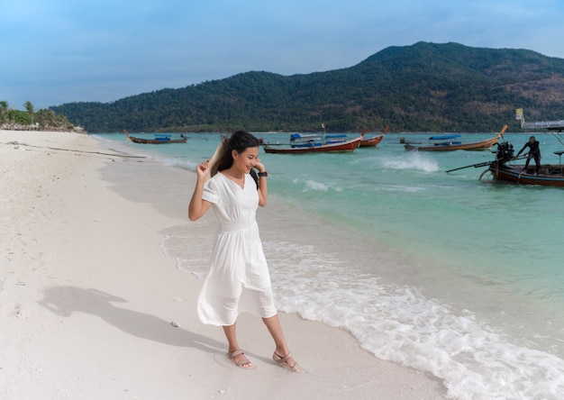 Asian woman in white dress holding hat happy smiling while playing with waves on blue sea beach