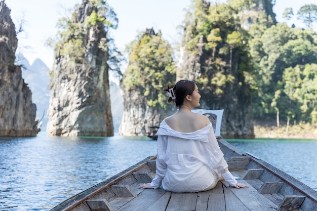 An Asian woman in a white shirt sits in front of a boat with a beautiful mountain in the middle of the sea during a trip in Thailand. Amzing thailand.