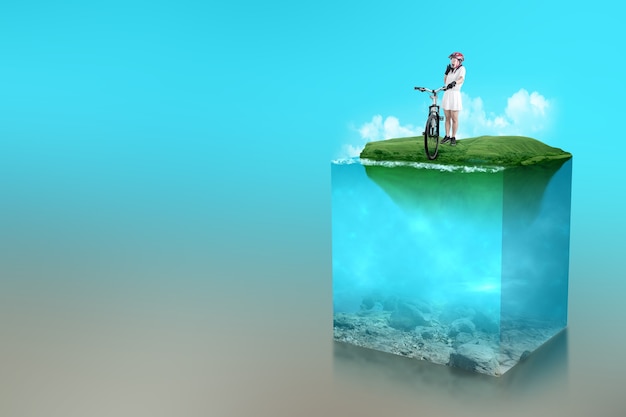 Asian woman with a bicycle helmet standing beside her bicycle on the field with an underwater view of blue water on the lake. Environment concept