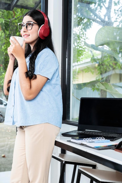 Asian woman with eyeglass listening to music with headphones and holding a cup of coffee with a laptop and a notebook with a mobile phone on the table