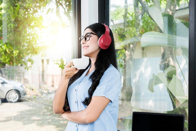 Asian woman with eyeglass listening to music with headphones and holding a cup of coffee