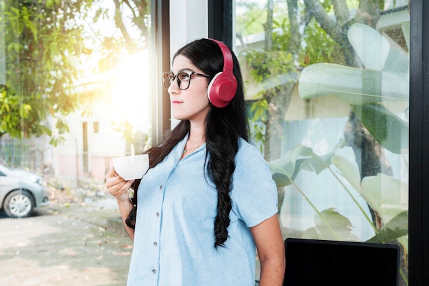 Asian woman with eyeglass listening to music with headphones and holding a cup of coffee