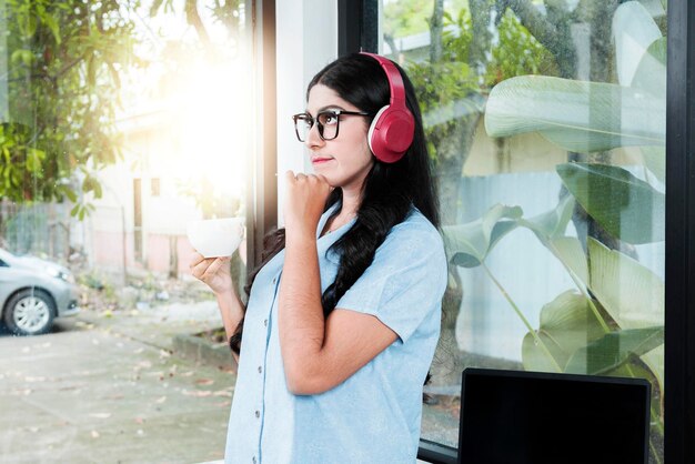 Asian woman with eyeglass listening to music with headphones and holding a cup of coffee