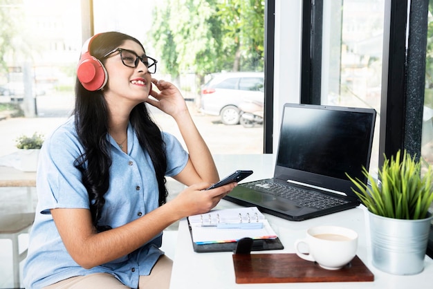 Asian woman with eyeglass using a mobile phone and headphones with a laptop and a notebook with a cup of coffee on the table