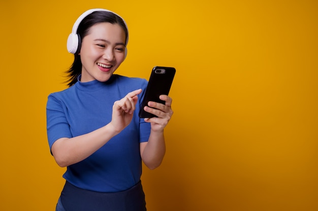 Asian woman with headphones listening to music from smart phone on yellow.