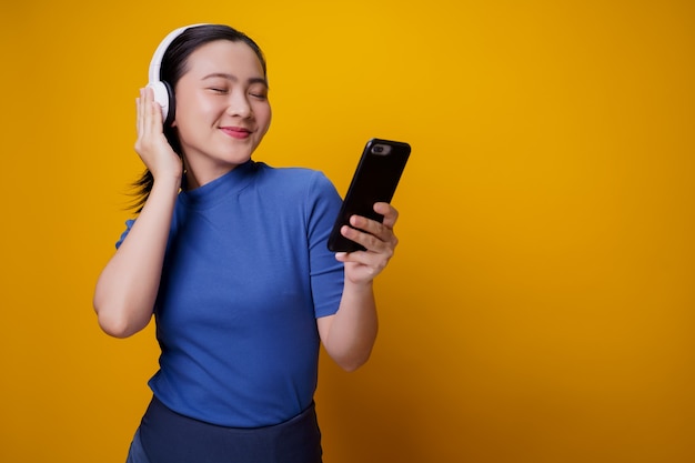 Asian woman with headphones listening to music from smart phone on yellow.