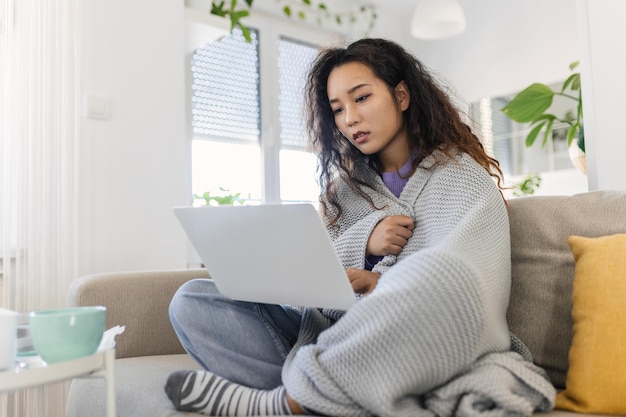 Asian woman wrapped in a cozy blanket sitting on the sofa and working on her laptop