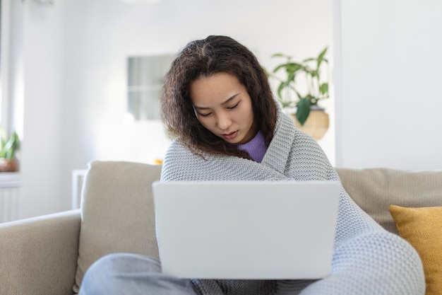 Asian woman wrapped in a cozy blanket sitting on the sofa and working on her laptop