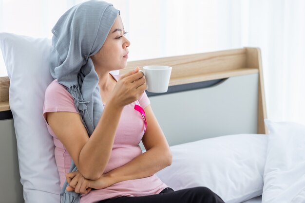 A asian women disease mammary cancer patient holding a coffee cup with pink ribbon wearing headscarf After treatment to chemotherapy on bed In the bedroom at the house,healthcare,medicine