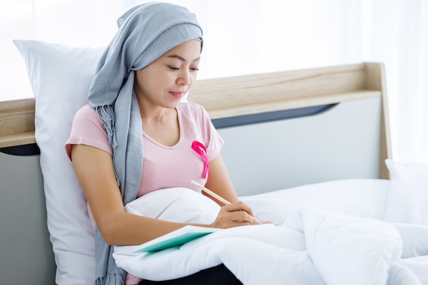 A asian women disease mammary cancer patient with pink ribbon wearing headscarf After treatment to chemotherapy with Take notes in a notebook on bed In the bedroom at the house,healthcare,medicine