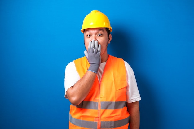 Asian worker wearing a helmet looks shocked hearing the news while closing his mouth with his hands 