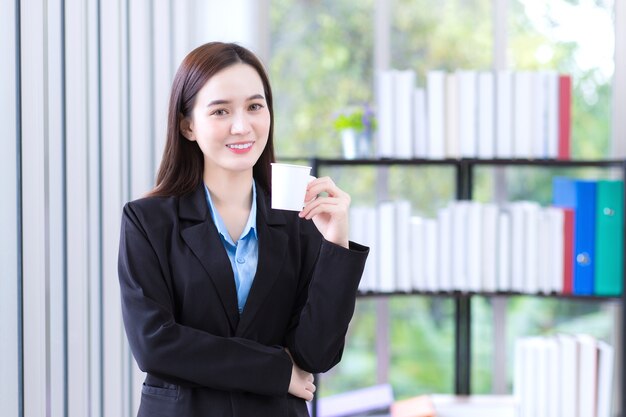 Asian working woman in a black suit holds paper cup to drink coffee in the morning at office.