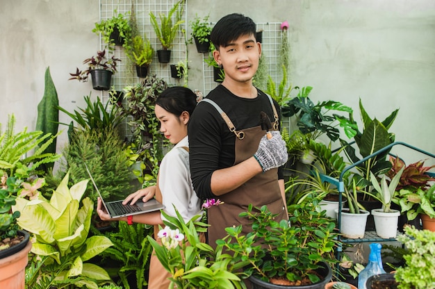 Asian young gardener couple wearing apron use garden equipment and laptop computer to research and take care the house plants in greenhouse