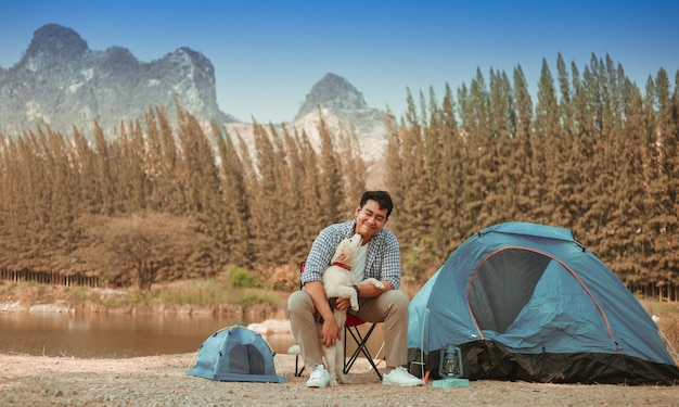 Asian young man in blue shirt with cute puppy dog camping on the lake hill mountain view happy and enjoy life