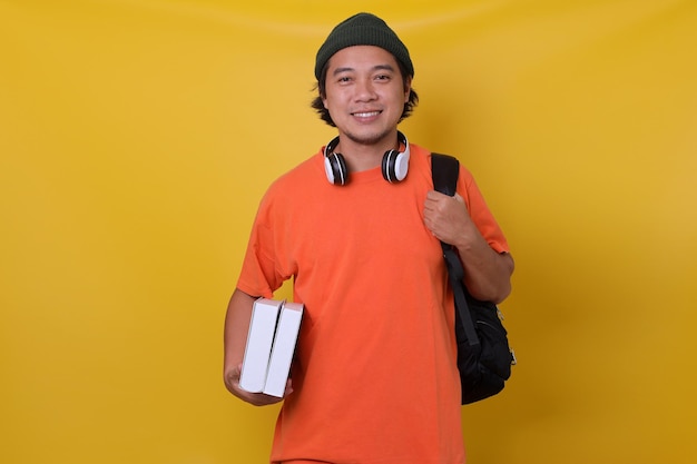 Photo asian young man in casual style with backpack and headphone bringing books isolated on yellow backgr