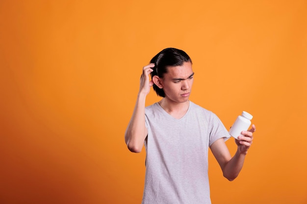 Asian young man confused about what pills to take, looking at tablets bottle. Disease treatment, illness remedy, upset puzzled patient holding medicaments, reading instruction