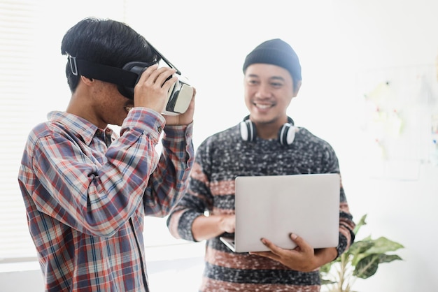 Asian young man controlling games using laptop with his friend wearing VR glasses.