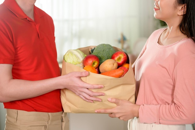 Asian young man delivery in red uniform holding healthy fruit vegetables in grocery bag send