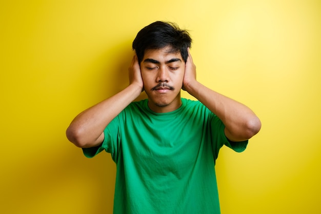 Asian young man in green t-shirt close his ears with both hands against yellow background