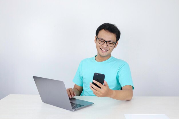 Asian young man happy and smile when he see on smart phone with laptop beside it Indonesian man wearing blue shirt