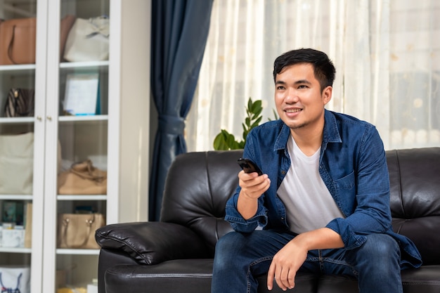 Asian young man holding remote control and watching TV while sitting on the couch at home