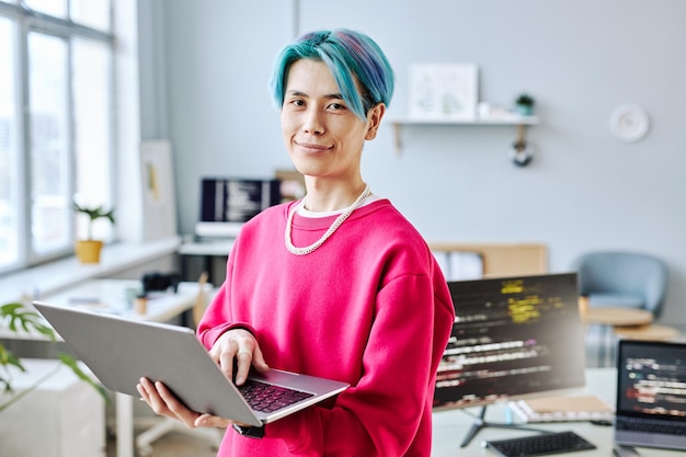 Asian young man looking at camera in office and holding laptop