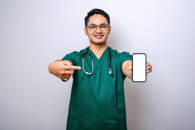 Asian young man nurse doctor wearing scrubs and stethoscope looking to camera showing blank smartphone screen recommending application