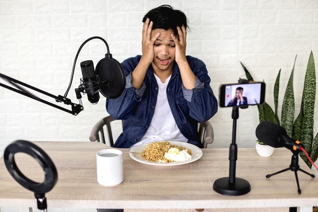 Asian young man surprised while seeing noodles on the dish during recording food vlog on smartphone.