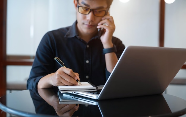 Asian young man talk mobile phones and take notes on their notebooks while working on laptop computers online business work from cafe teleworking concept