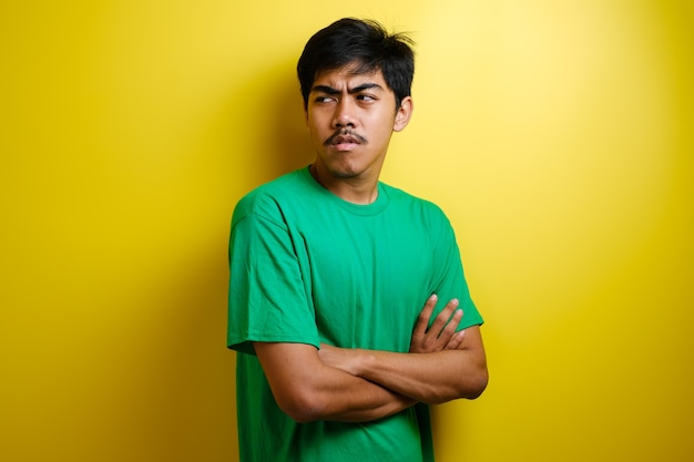 Asian young man wearing yellow t-shirt crossed his arm on chest while looking to his side in anger. Yellow background