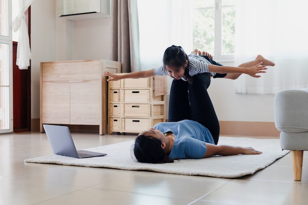 Asian young mother and her daughter doing stretching fitness exercise yoga together at home. Parent teaching child work out to be strong and maintain physical health and wellbeing in daily routine.