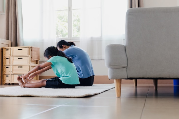 Asian young mother and her daughter doing stretching fitness exercise yoga together at home. Parent teaching child work out to be strong and maintain physical health and wellbeing in daily routine.