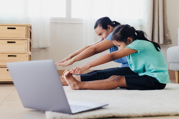 Asian young mother and her daughter doing stretching fitness exercise yoga together at home. Parent teaching child work out to be strong and maintain physical health and wellbeing in daily routine.