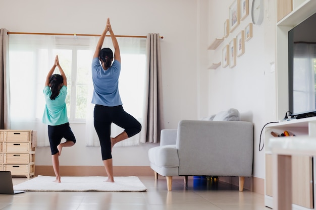 Asian young mother and her daughter doing stretching fitness exercise yoga together at home. Parent teaching child work out to be strong and maintain physical health and wellbeing in daily routine.