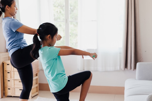 Asian young mother and her daughter doing stretching fitness exercise yoga and using the bottle of water to be dumbbells together at home in daily routine.