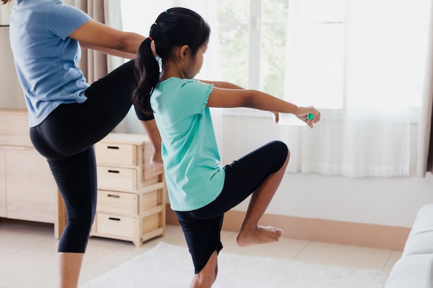 Asian young mother and her daughter doing stretching fitness exercise yoga and using the bottle of water to be dumbbells together at home in daily routine.