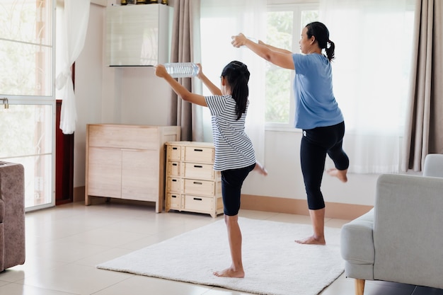 Asian young mother and her daughter doing stretching fitness exercise yoga and using the bottle of water to be dumbbells together at home in daily routine.