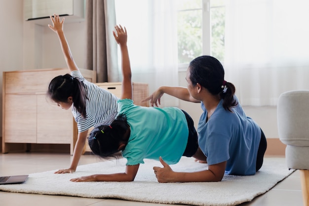 Asian young mother and her daughters doing stretching fitness exercise yoga together at home. Parent and children work out to be strong and maintain physical health and wellbeing in daily routine.