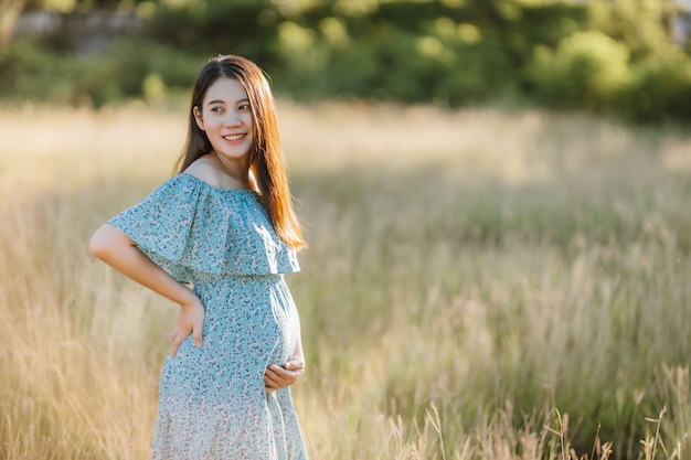 Asian young pregnant woman in blue dress relaxing and enjoying life in grass nature at summer day.