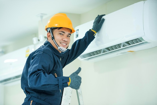 An Asian young Technician service man wearing blue uniform checkingProfessional air conditioner installer maintaining modern indoor air conditioner space for text