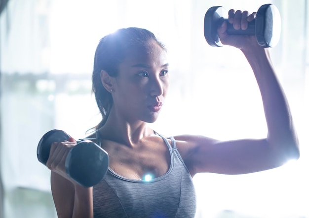 Asian young woman doing dumbbell workout at home working on arms strength athletic female lifting blue fitness dumbells up over living room interior